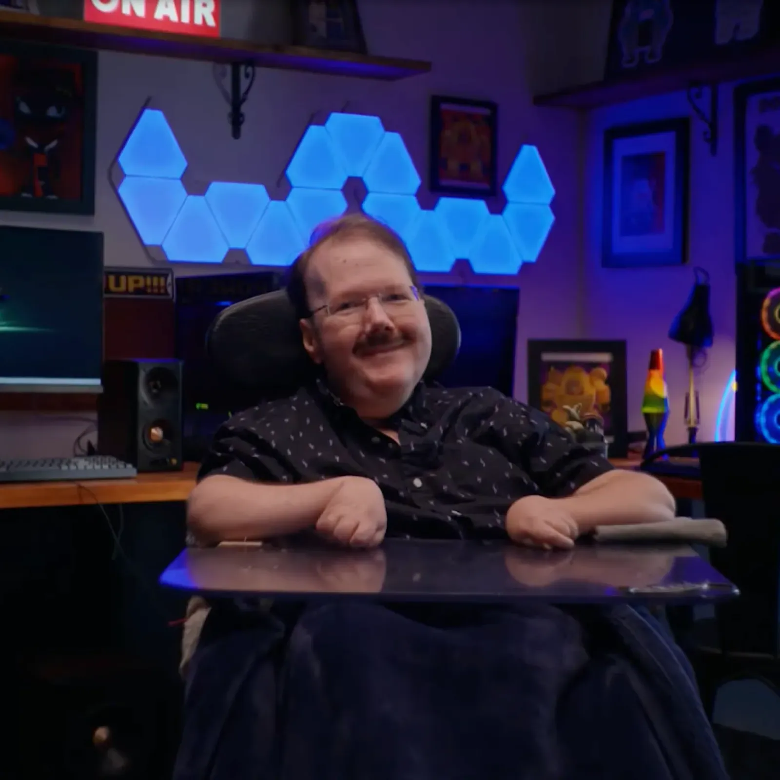 A smiling white man with muscular dystrophy sits at his motorized wheelchair, in front of a monitor gaming setup and blue-lit desk